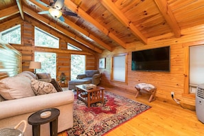 A log cabin interior with a sofa, armchairs, a TV, and a large window.