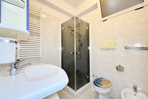 Une salle de bain dotée d’une douche à parois de verre, d’un lavabo blanc, de toilettes avec un abattant coloré et d’un miroir au-dessus du lavabo.