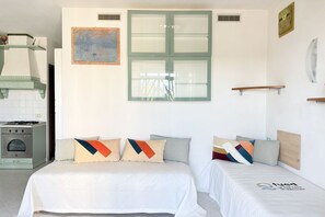 Une chambre d’hôtel avec deux lits, un coin cuisine, un tableau et une fenêtre avec des stores.