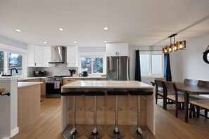 A modern kitchen with a central island, bar stools, a dining area, and a large refrigerator. - Luxe 3BR • Sleeps 8 • King Beds • Steps to Train (Calgary)