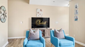 A living room with two blue armchairs, a flat-screen TV, a clock, and wall art. - Horizon South 55-304 (Panama City Beach)