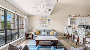 A spacious living area with a large window, a ceiling fan, a sofa with cushions, a coffee table, and a kitchen area with bar stools. - Horizon South 55-304 (Panama City Beach)