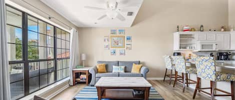 A spacious living area with a large window, a ceiling fan, a sofa with cushions, a coffee table, and a kitchen area with bar stools.