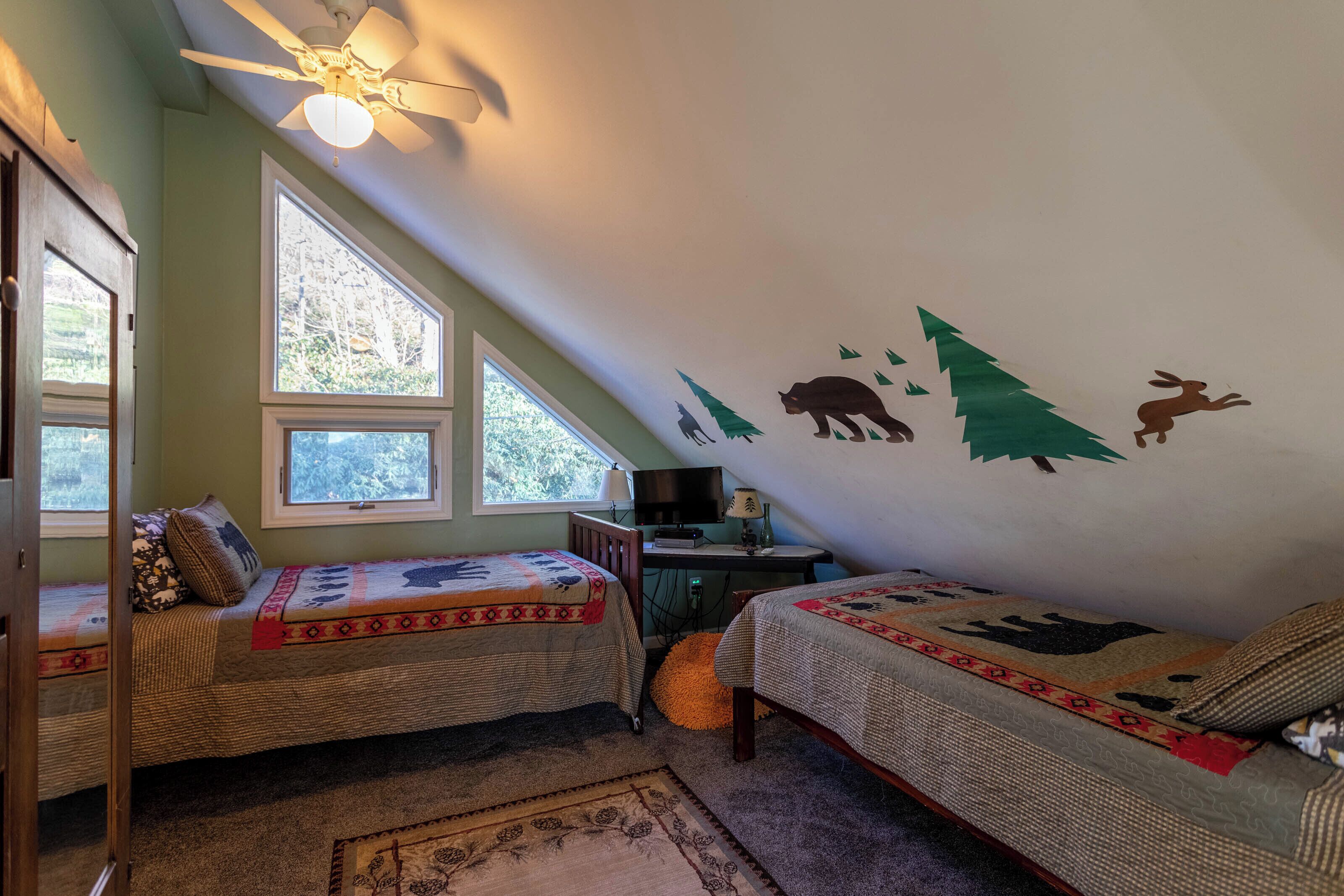 Une chambre mansardée chaleureuse avec deux lits superposés, un ventilateur de plafond, un bureau avec un ordinateur et une grande fenêtre donnant sur les arbres.