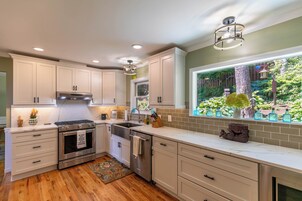 Una cocina moderna con armarios blancos, electrodomésticos de acero inoxidable, un amplio ventanal con vistas a zonas verdes y un jarrón decorativo en la encimera.