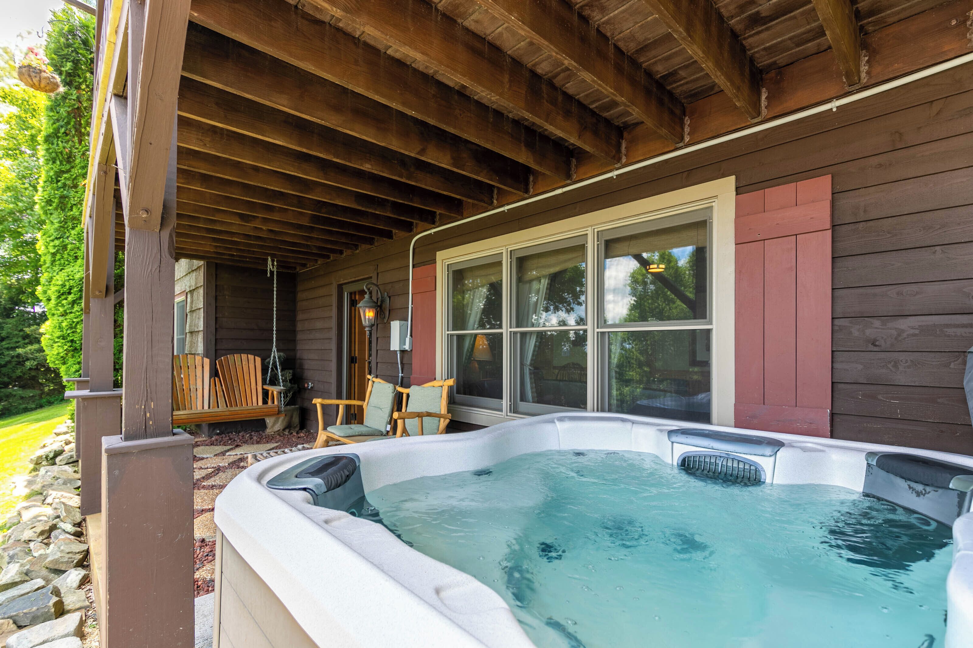 A hot tub under a wooden pergola with a stone wall and chairs outside.
