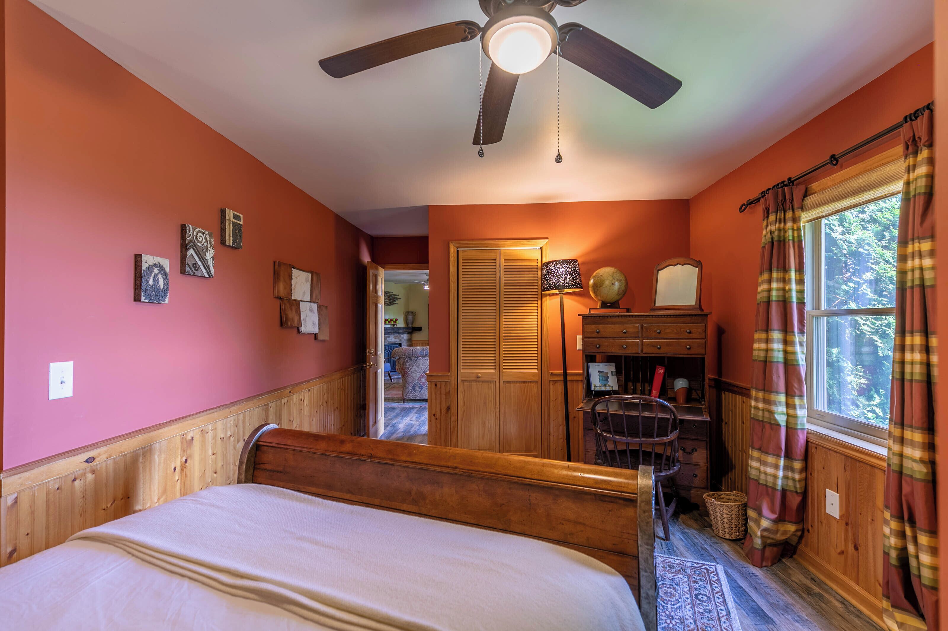 A bedroom with a bed, wooden headboard, ceiling fan, wardrobe, desk, chair, window with curtains, and a view of the outside.