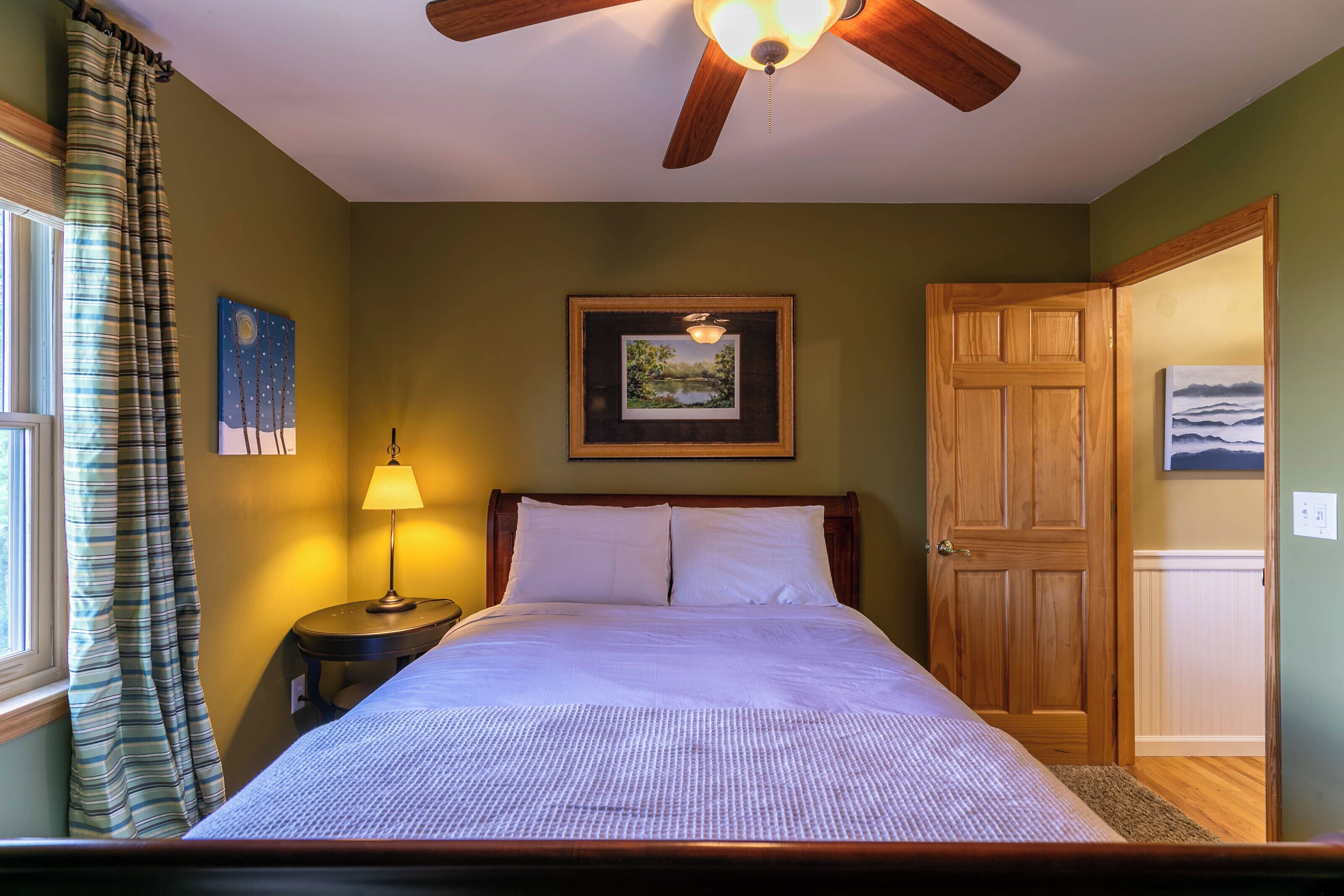Une chambre à coucher avec un lit, une table de chevet, une lampe, un ventilateur de plafond, une porte et un tableau encadré au mur.