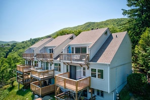 A multi-story cabin with a deck, surrounded by forested hills.