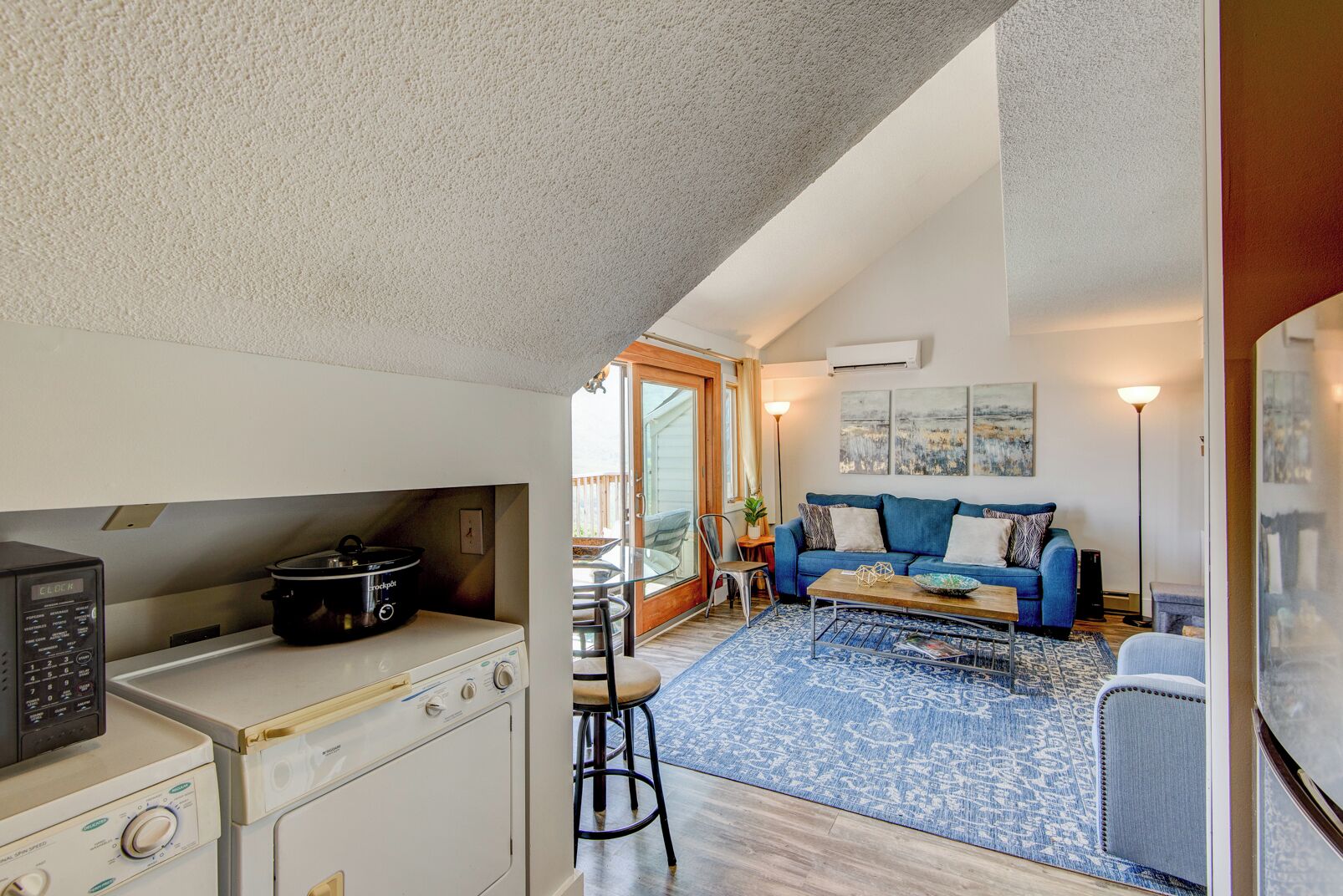 A compact kitchen with a stove, a slow cooker, and a washing machine. There is a blue sofa with cushions and a coffee table in the living area.