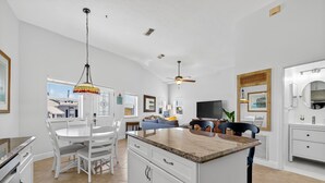 A modern kitchen with white cabinetry, a dining area with a white table and chairs, a ceiling fan, and a TV. - Beachbound 2BR Apt 1282 (Jensen Beach)