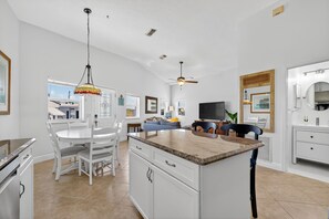 Eine moderne Küche mit weißen Schränken, ein Essbereich mit weißem Tisch und Stühlen, ein Deckenventilator und ein Fernseher.