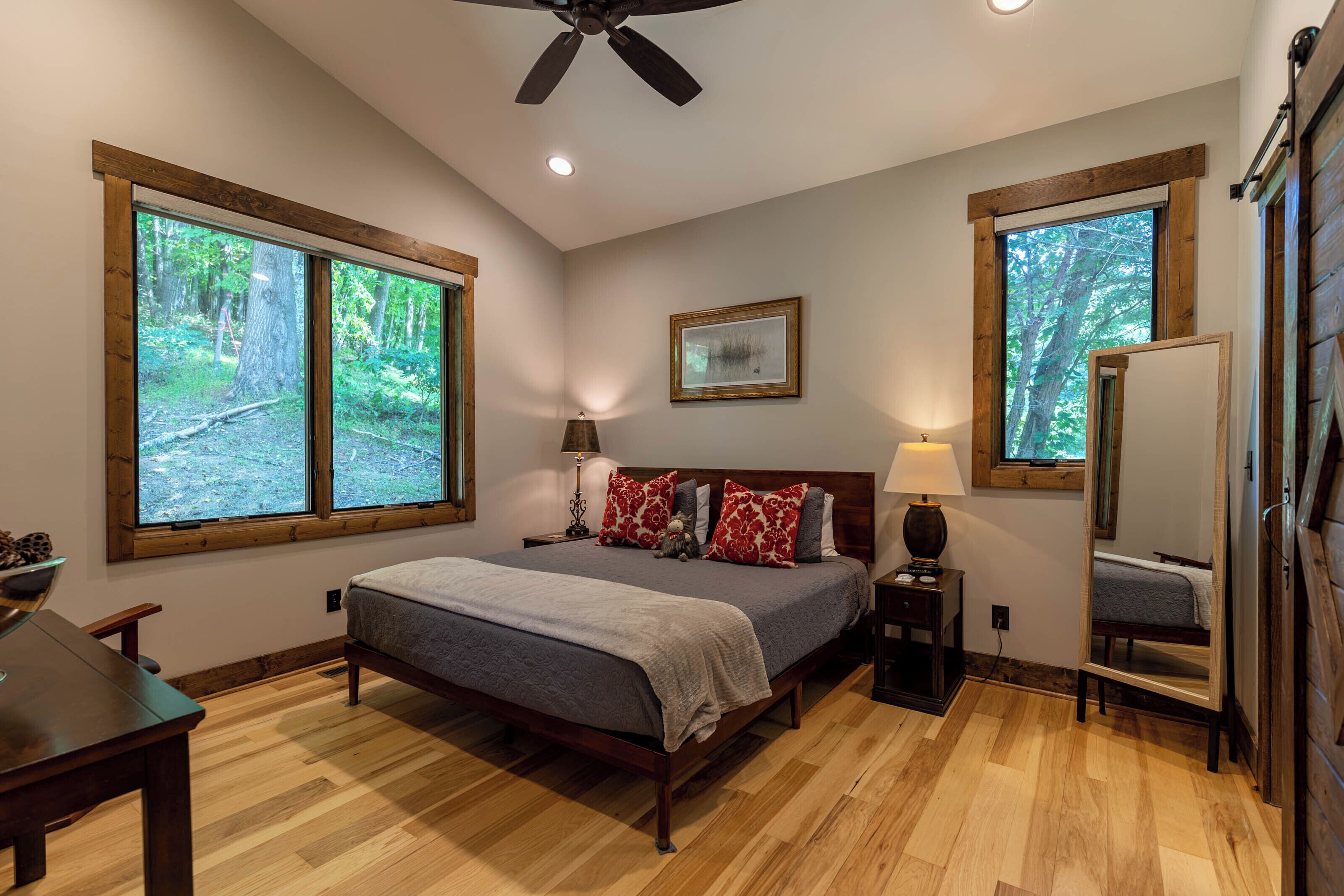 A bedroom with a large bed, two windows with forest views, a ceiling fan, and a bedside table with a lamp.