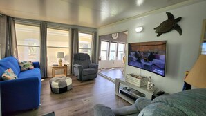 A living room with a blue sofa, grey armchair, and ottoman. A television is mounted on the wall displaying a news program. - Isle Always Luv View 2BR 1277 (Jensen Beach)