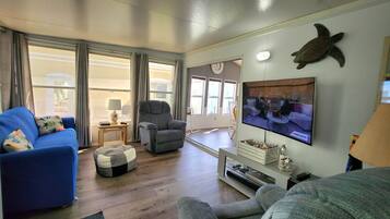 Ein Wohnzimmer mit einem blauen Sofa, einem grauen Sessel und einem Hocker. Ein Fernseher an der Wand zeigt eine Nachrichtensendung.