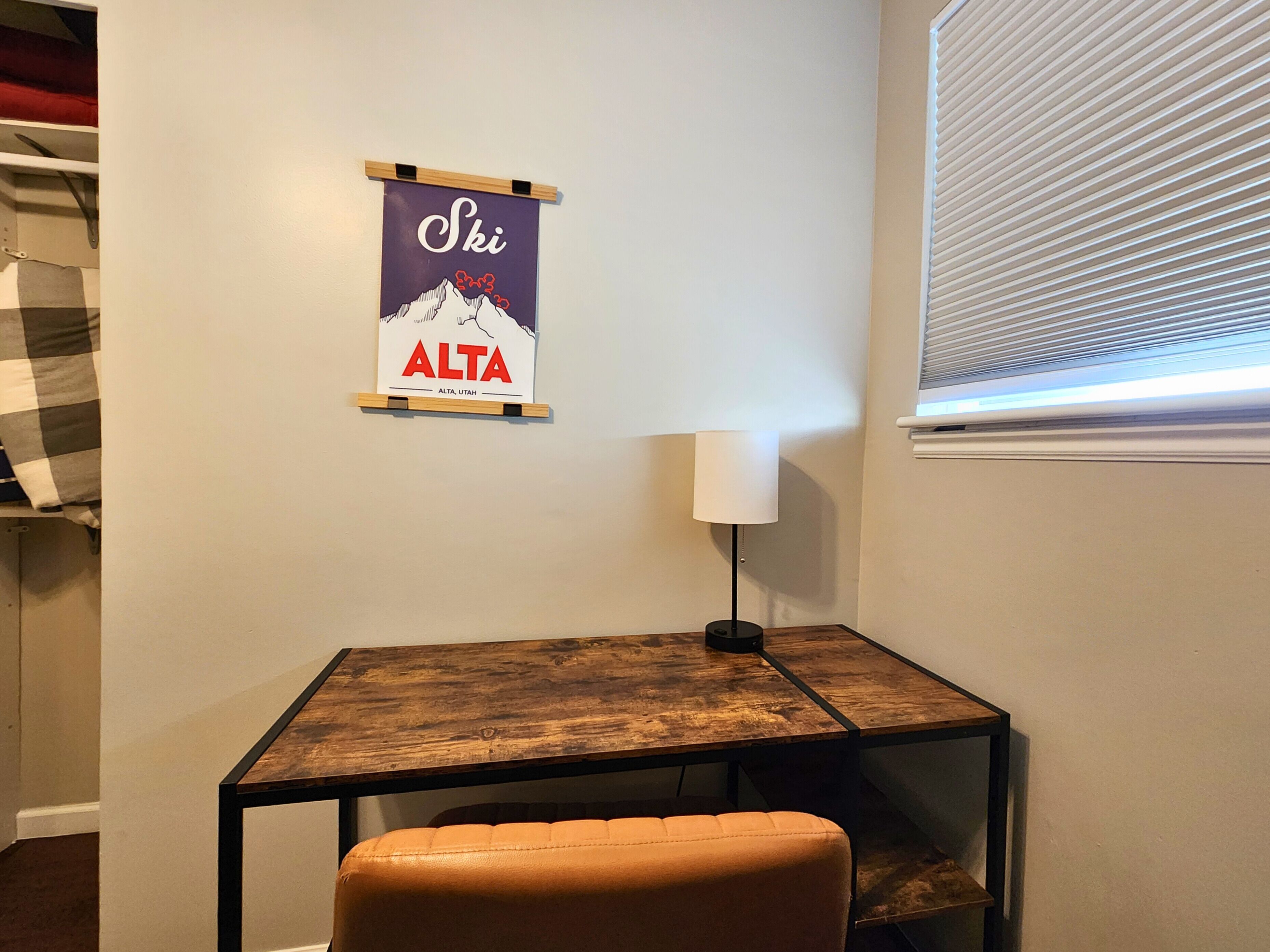 A small room with a wooden table, a single chair, a wall-mounted lamp, and a poster of a mountain.