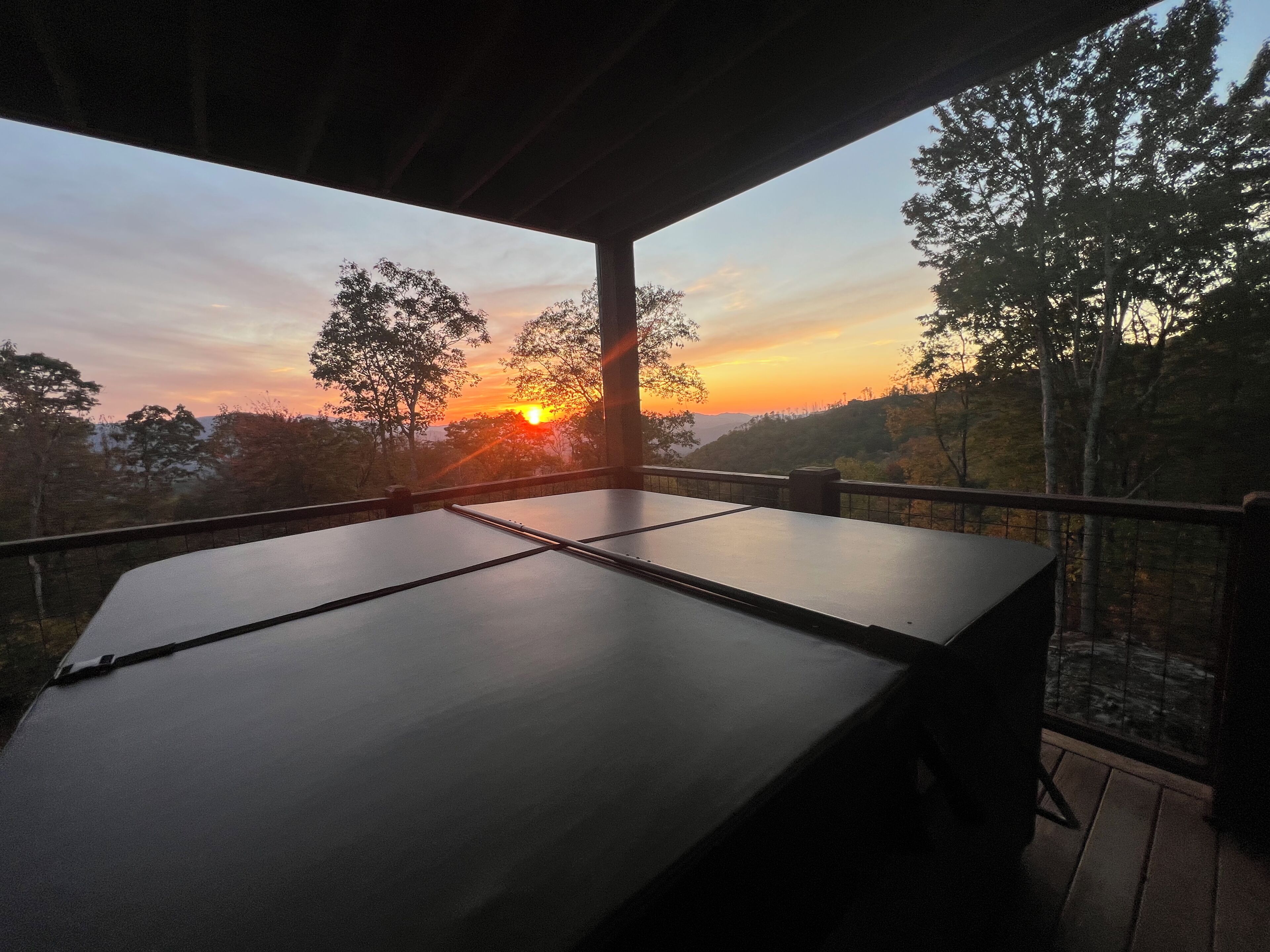 A covered outdoor area with a table and chairs, overlooking a sunset with trees and hills in the background.