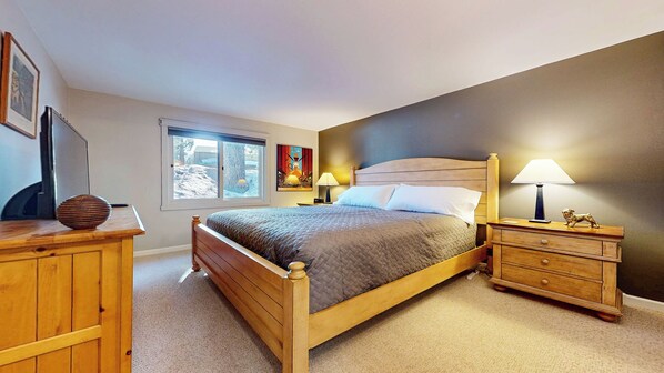 A bedroom with a wooden bed, a nightstand with a lamp, a dresser, a window with a view of trees, and a painting on the wall.