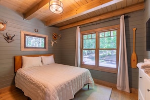 A bedroom with a bed, a window with curtains, a wooden ceiling, and wall-mounted deer antlers.