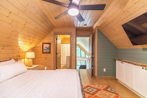 A bedroom with a bed, a ceiling fan, a nightstand, a lamp, a window, and a view of the outside.