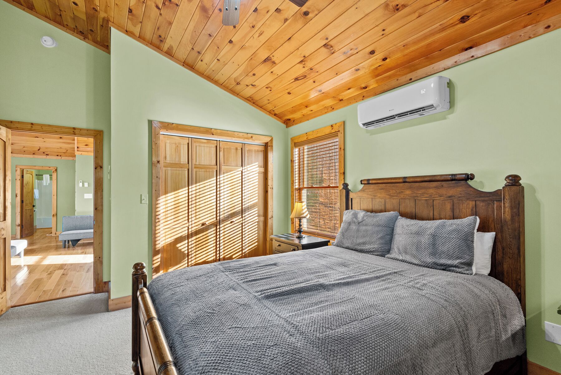 Un dormitorio con techo de madera, una cama con manta tejida color gris, cabecera de madera y una ventana con persianas.