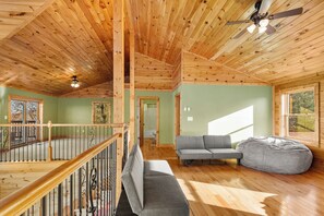 Ein geräumiges Zimmer mit Holzbalkendecke, einem grauen Sofa, einem Sitzsack und einem Balkon mit Geländer.