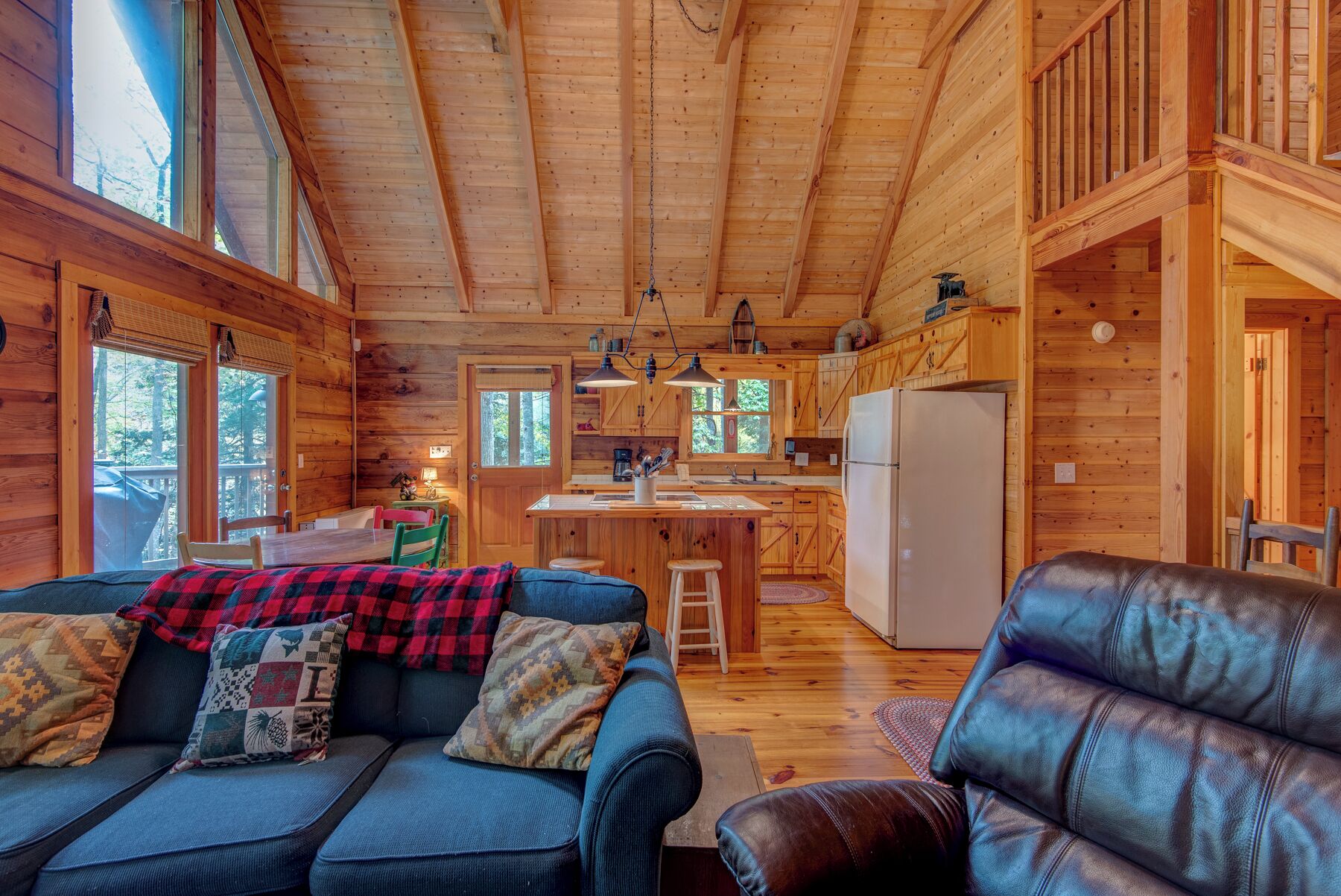 A cozy cabin interior with a wooden ceiling, a dining area, a kitchen, and a living room with a sofa and a leather chair.