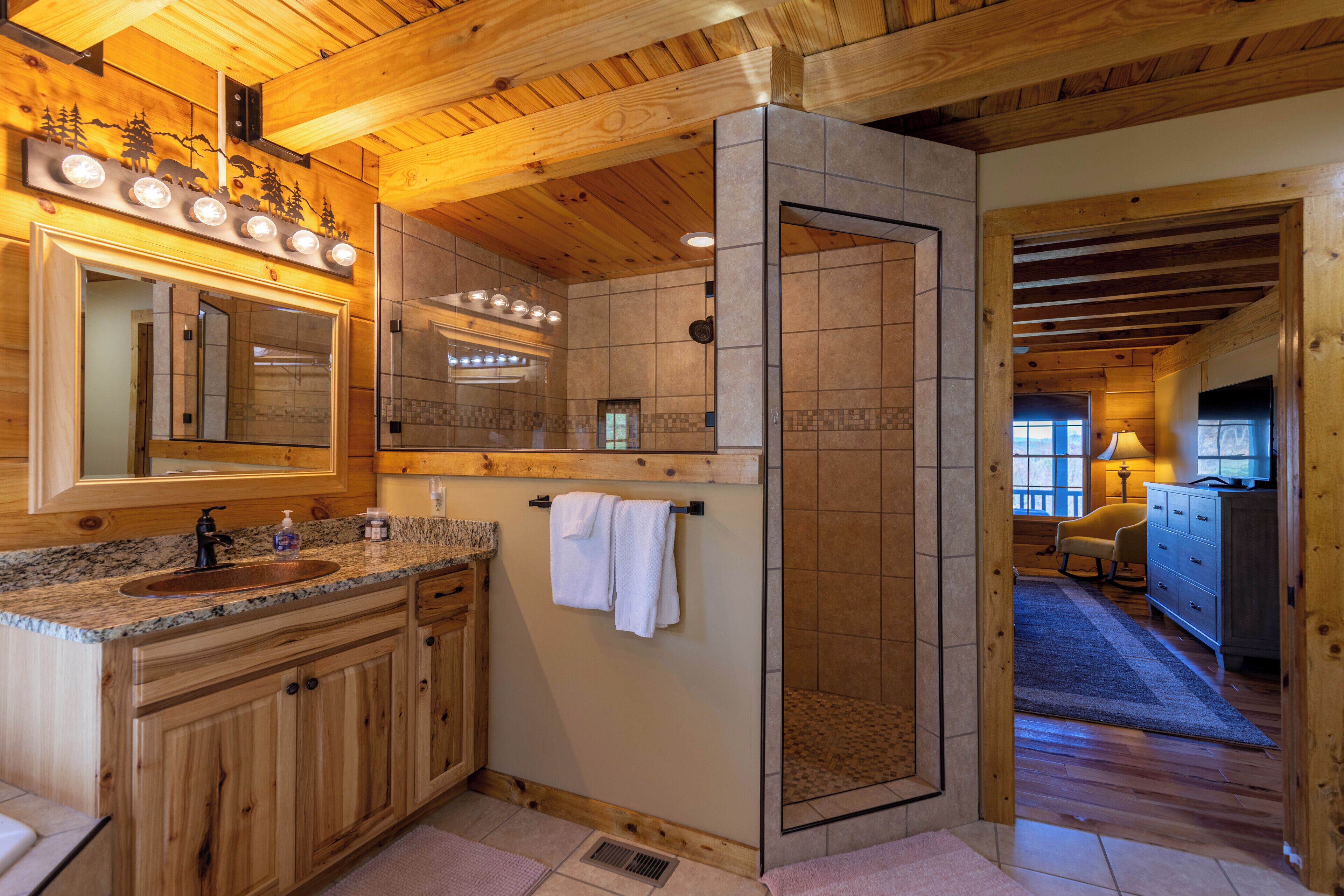 A wooden cabin interior with a bathroom featuring a granite countertop sink, wooden cabinets, a large mirror, and a shower area.
