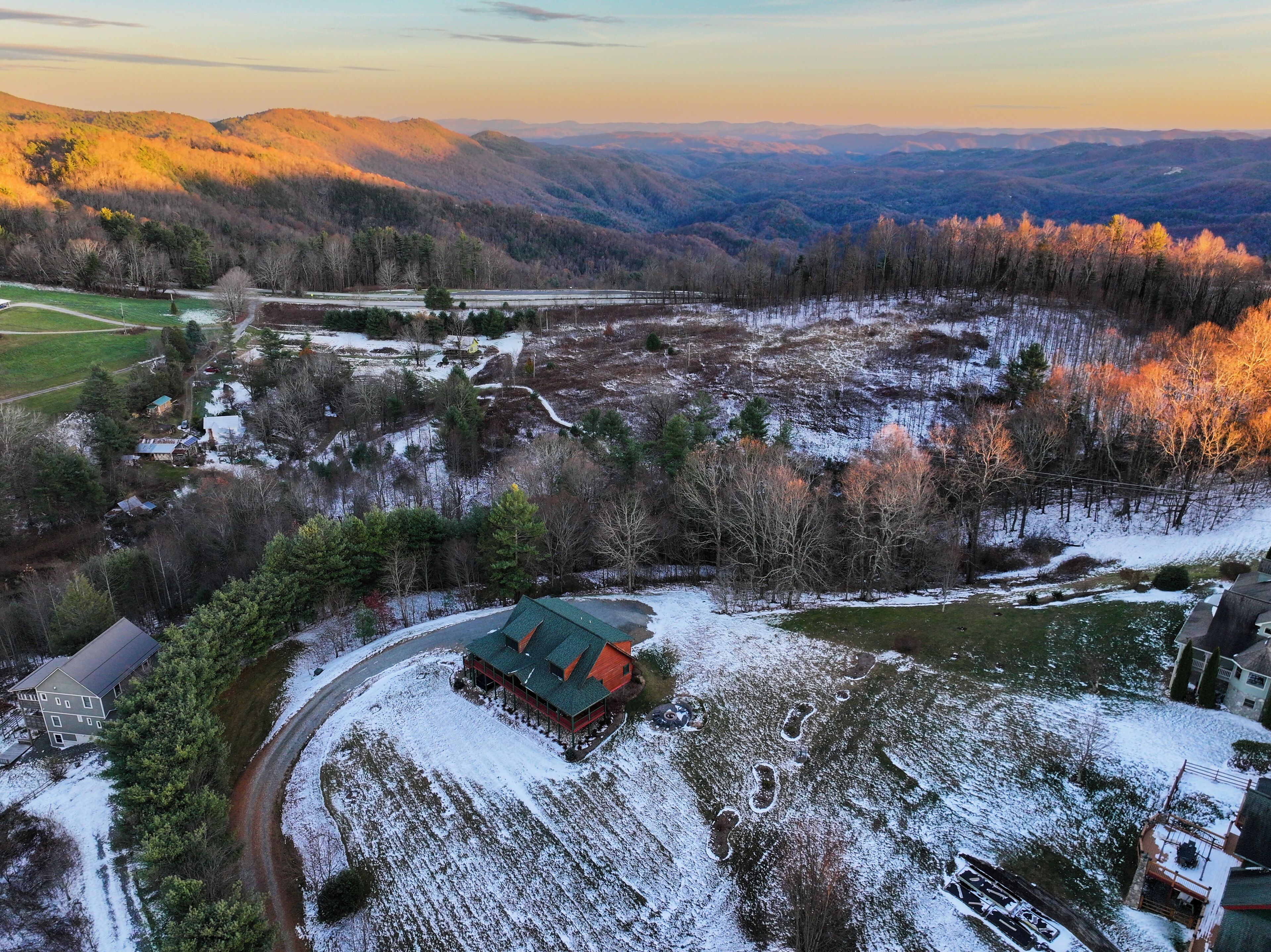 A rural property with a large house featuring a green roof, surrounded by snow-covered fields and forested hills.