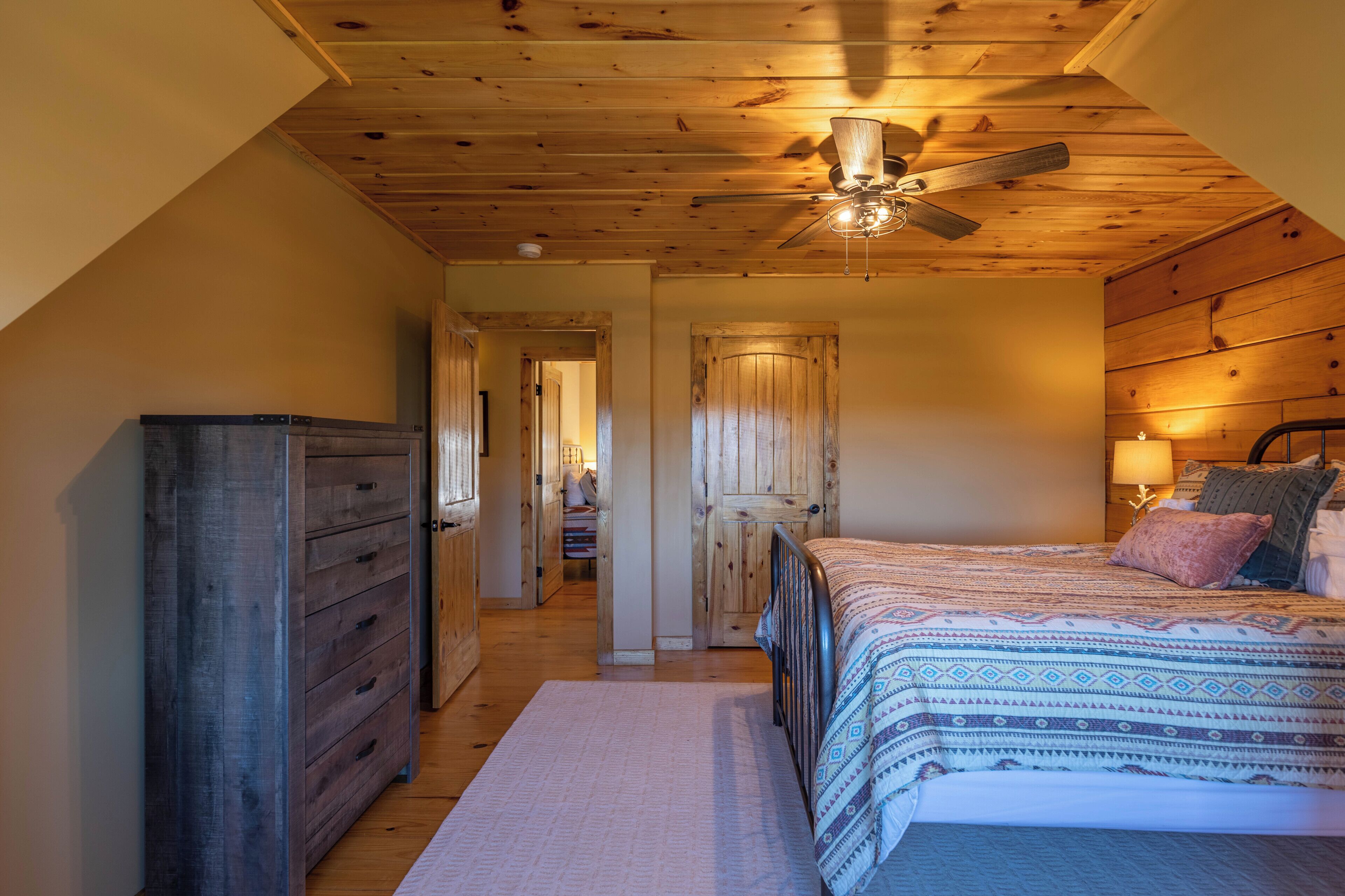 Une chambre avec un plafond en bois, un lit recouvert d’une literie à motifs, une commode et un ventilateur de plafond.