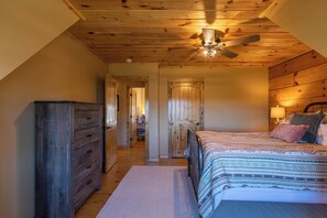 Ein Schlafzimmer mit Holzbalkendecke, einem Bett mit gemusterten Betttextilien, einem Schminktisch und einem Deckenventilator.