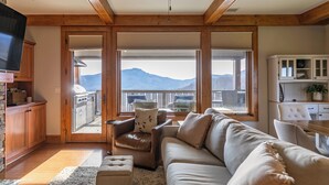 A living room with a sofa, armchair, and TV, a balcony with a view of mountains, and wooden beams on the ceiling. - Endless Grandfather View at Echota (Boone)