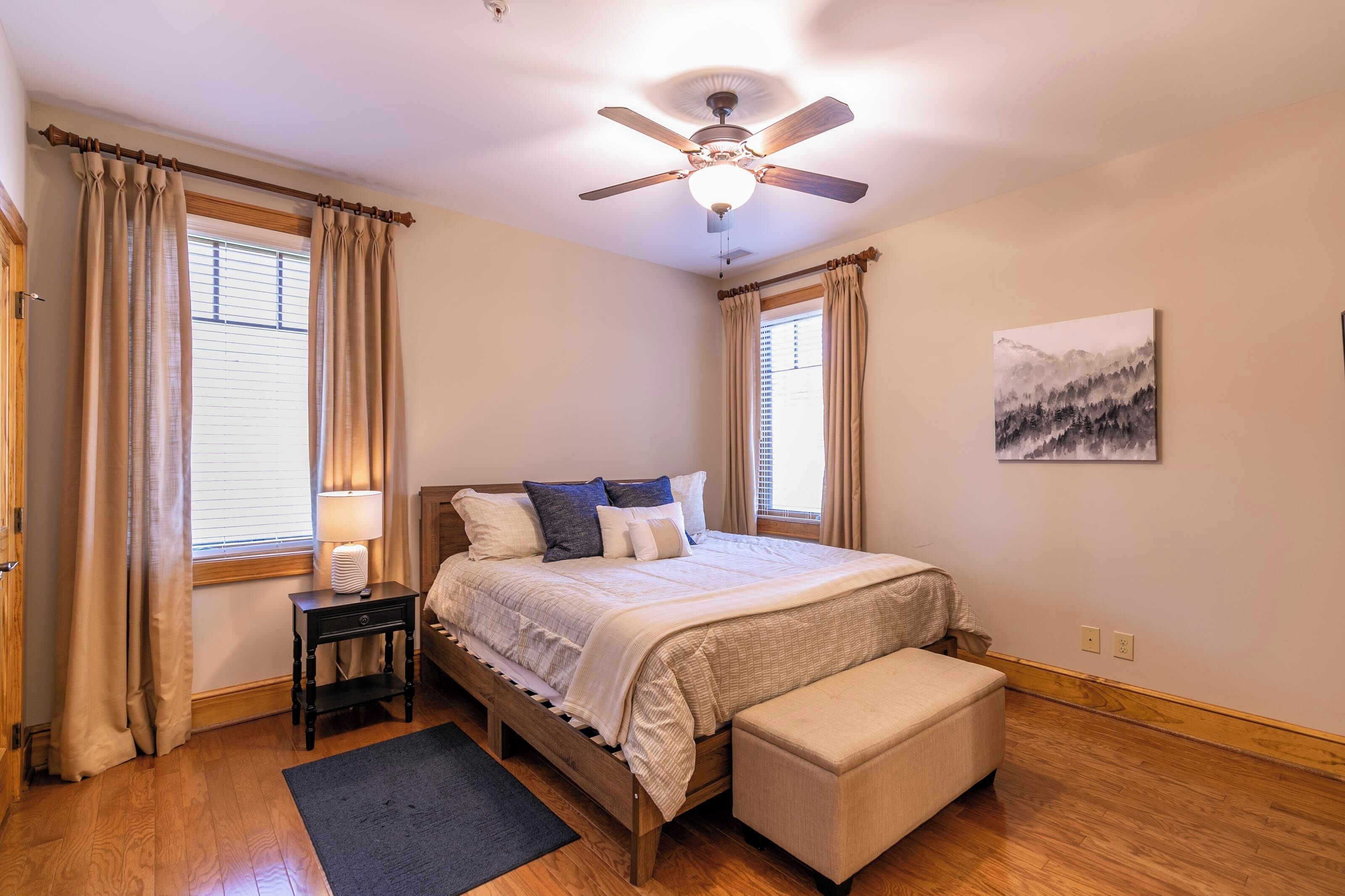 A bedroom with a bed, bedside table, bench, ceiling fan, and artwork on the wall.