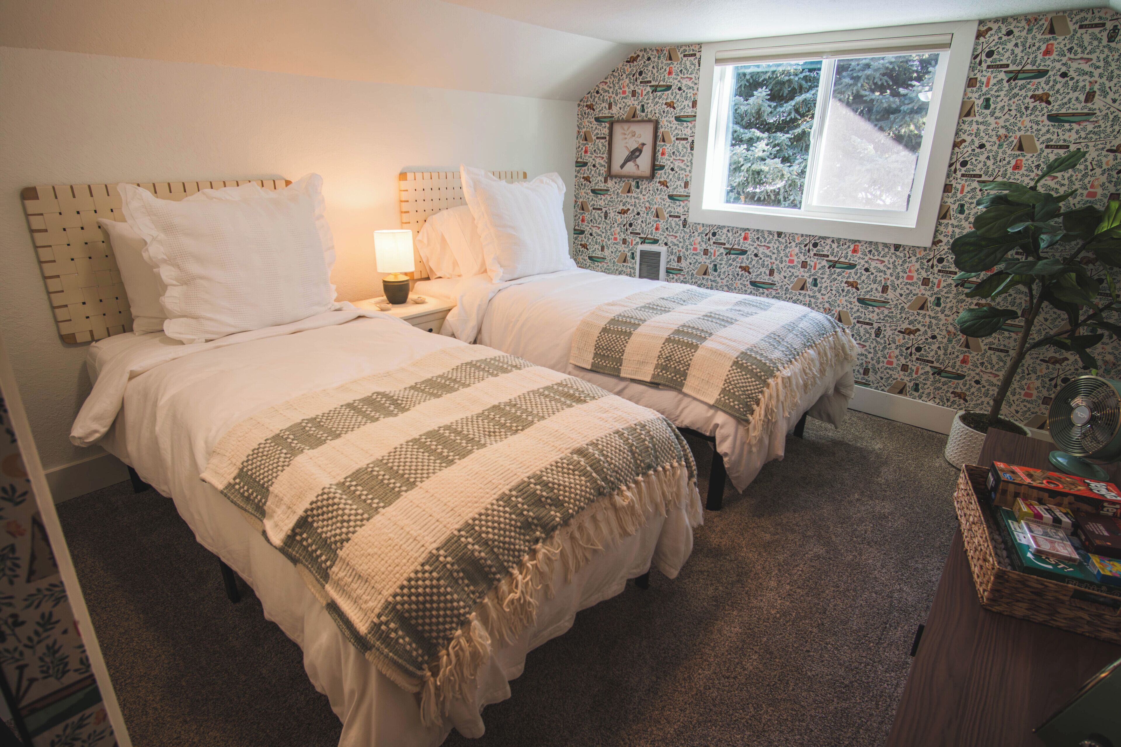 A bedroom with two beds, a window, a plant, and a bedside table with books.