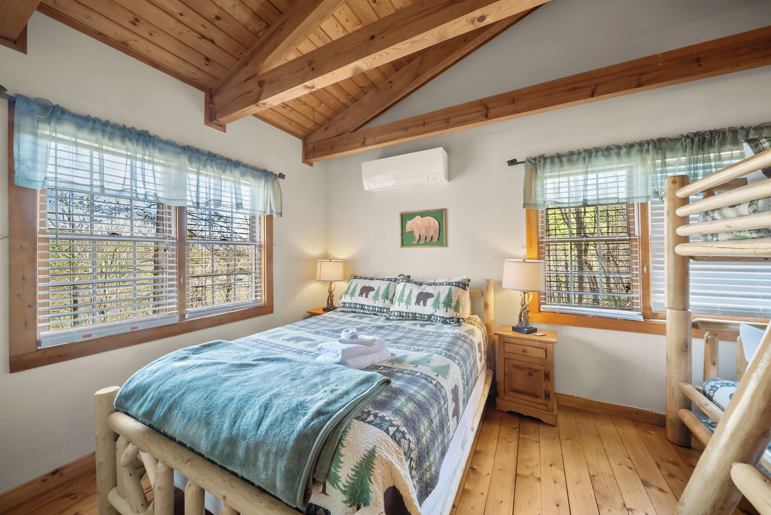 A wooden-framed bedroom with a bed, two windows with blinds, a wall-mounted air conditioner, a nightstand with a lamp, and a chair.