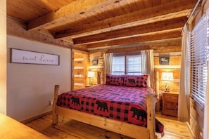 A cozy log cabin bedroom with a bed, wooden walls, a window with blinds, a bedside table with a lamp, and a framed picture on the wall. - 5 Bears Cabin Wooded Cabin With hot Tub, Arcade Games, Pool Table, Fire pit (Banner Elk)