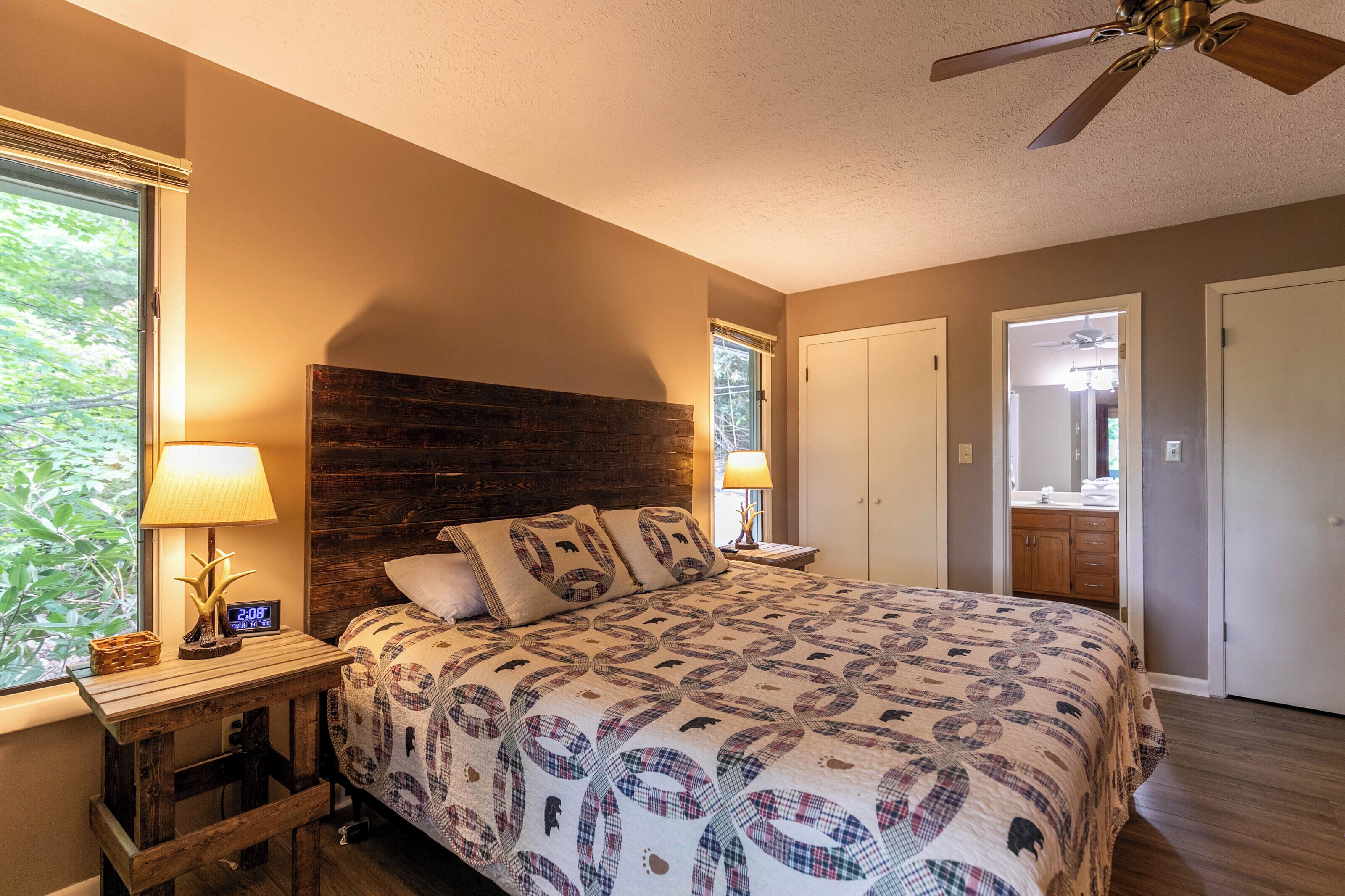A bedroom with a bed, a wooden headboard, a window with a view, a ceiling fan, and a nightstand with a lamp.
