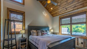 A bedroom with a wooden ceiling, a bed with a headboard, a nightstand, a lamp, and a window with a view of greenery. - Happy Camper at Eagles Nest (Banner Elk)