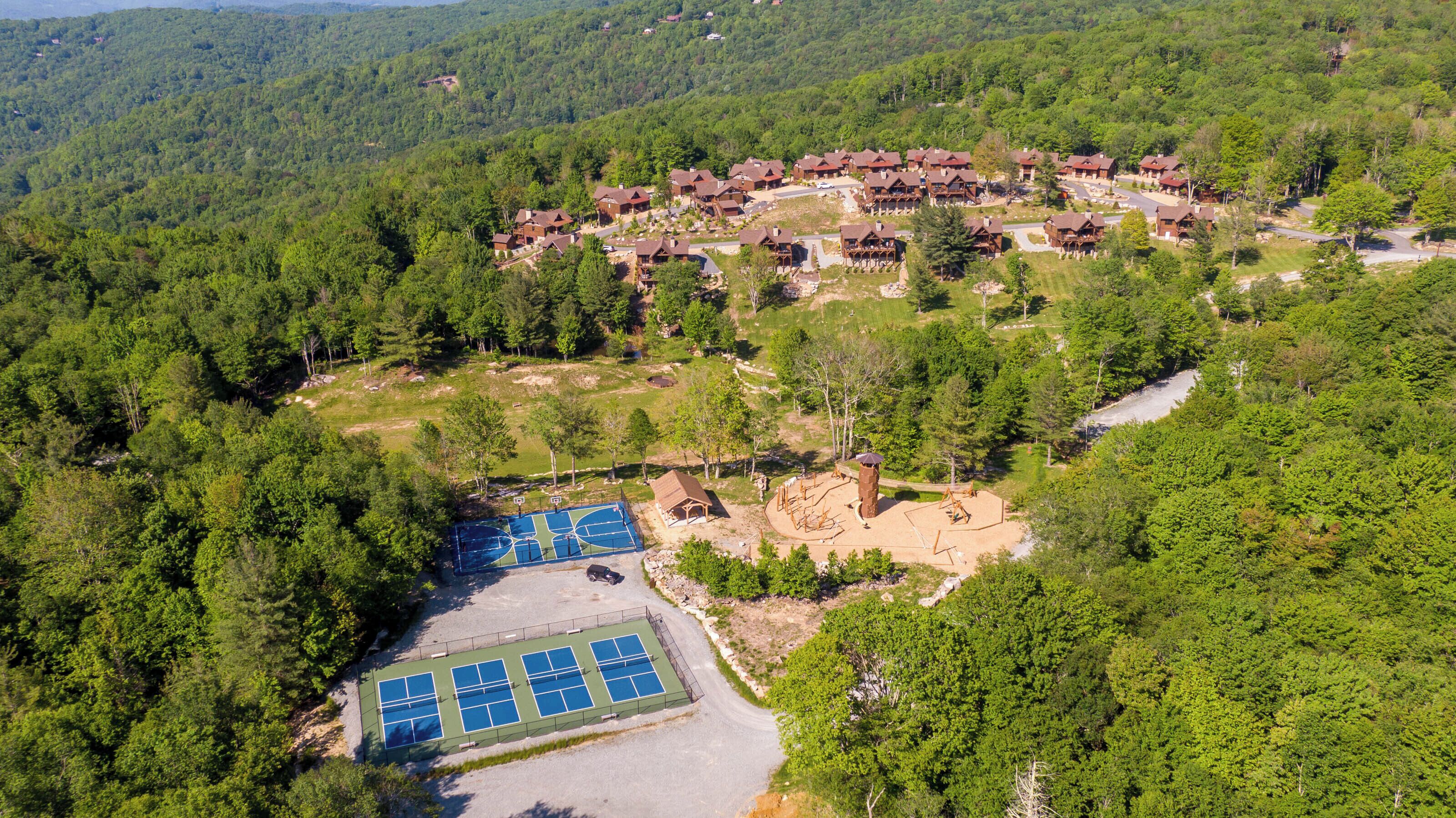 Vista aérea de un complejo turístico con varias cabañas de madera, una cancha de tenis y un parque infantil.