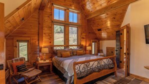 Een slaapkamer in een blokhut met een groot bed, een stoel, een nachtkastje en een raam met uitzicht op een bos.