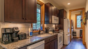 A kitchen with dark wooden cabinets, a granite countertop, a microwave, a refrigerator, a dishwasher, and a window with a view of trees. - Carolina Wren Cottage (Blowing Rock)