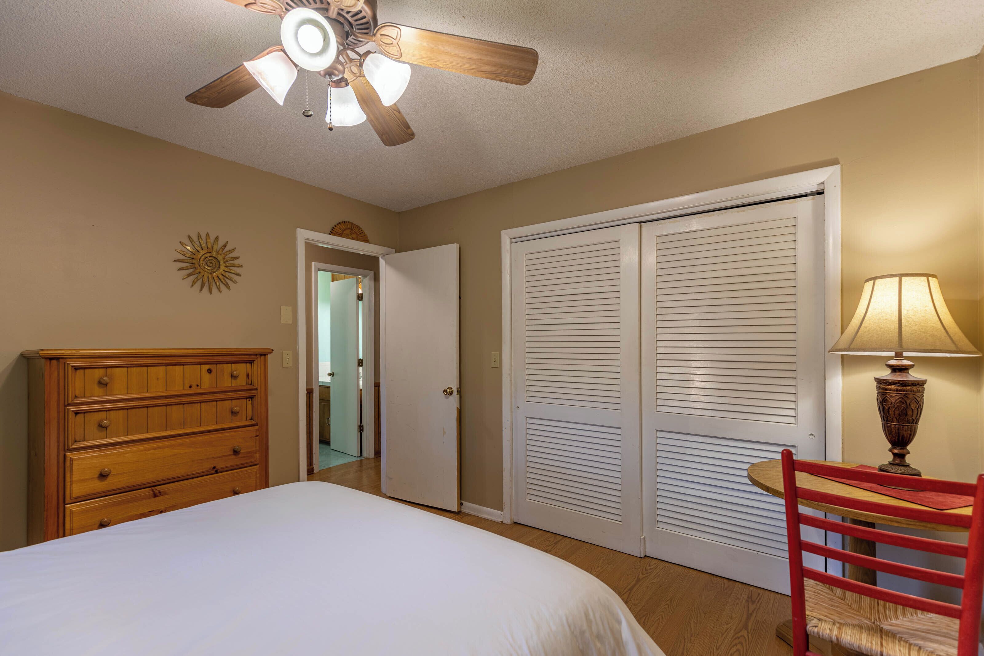 A bedroom with a bed, a wooden dresser, a ceiling fan, a door, and a table with a lamp.