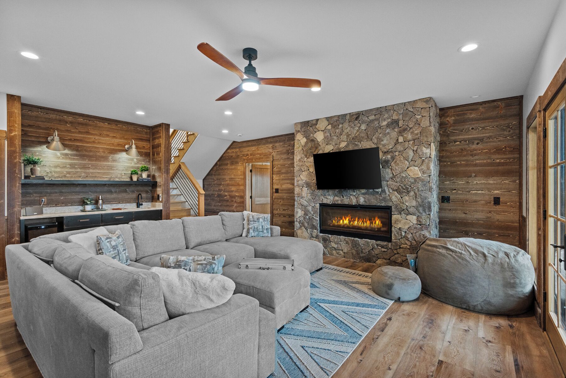 Ein modernes Wohnzimmer mit Ecksofa, Kamin aus Stein, Deckenventilator und Küchenbereich mit Holzdetails.