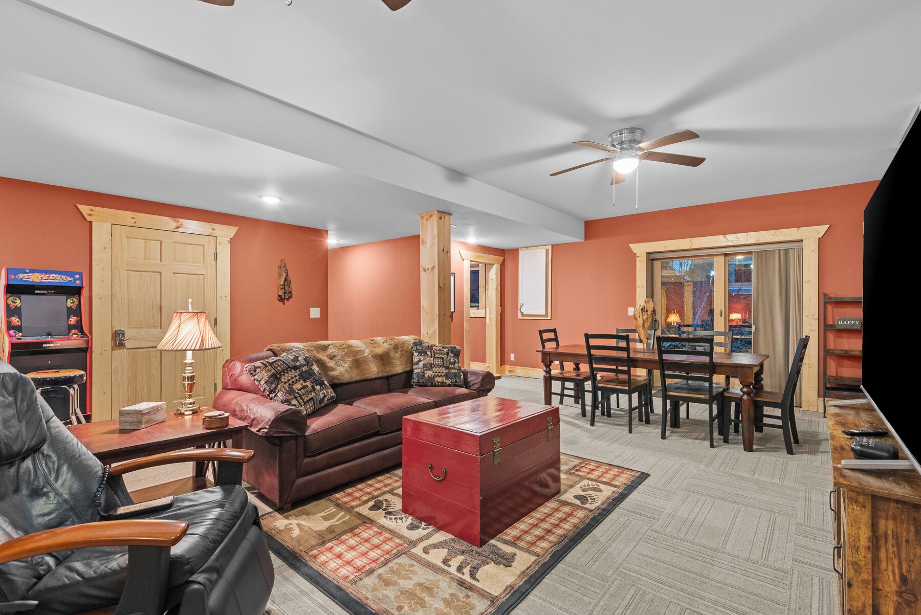 A living room with a sofa, a red chest of drawers, a dining table with chairs, a ceiling fan, and a wooden door.