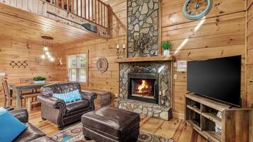 A log cabin interior with a stone fireplace, leather furniture, a wooden dining area, and a mounted clock.