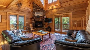 A cozy cabin interior with a stone fireplace, leather sofas, a wooden coffee table, and a TV mounted on the wall. - Morris Cabin (Boone)