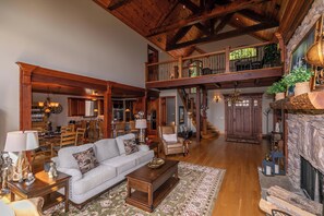 A spacious living area with a high ceiling, wooden beams, a fireplace, and a dining area with a kitchen in the background. - Haven at Headwaters Long-range Mountain Views, Pet Friendly With Game Room (Elk Park)