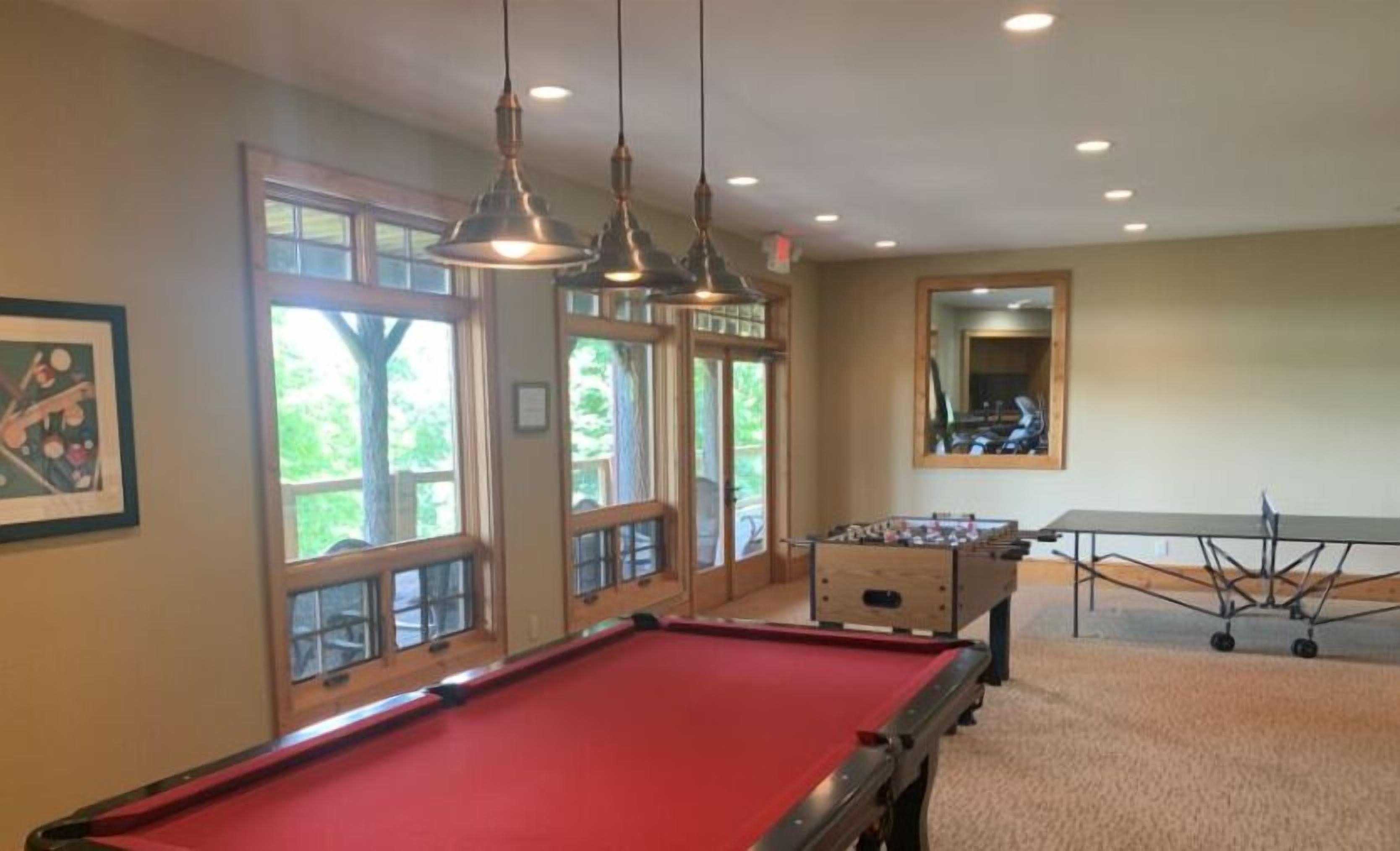 A pool table, a foosball table, and a billiards table in a room with large windows and hanging lights.