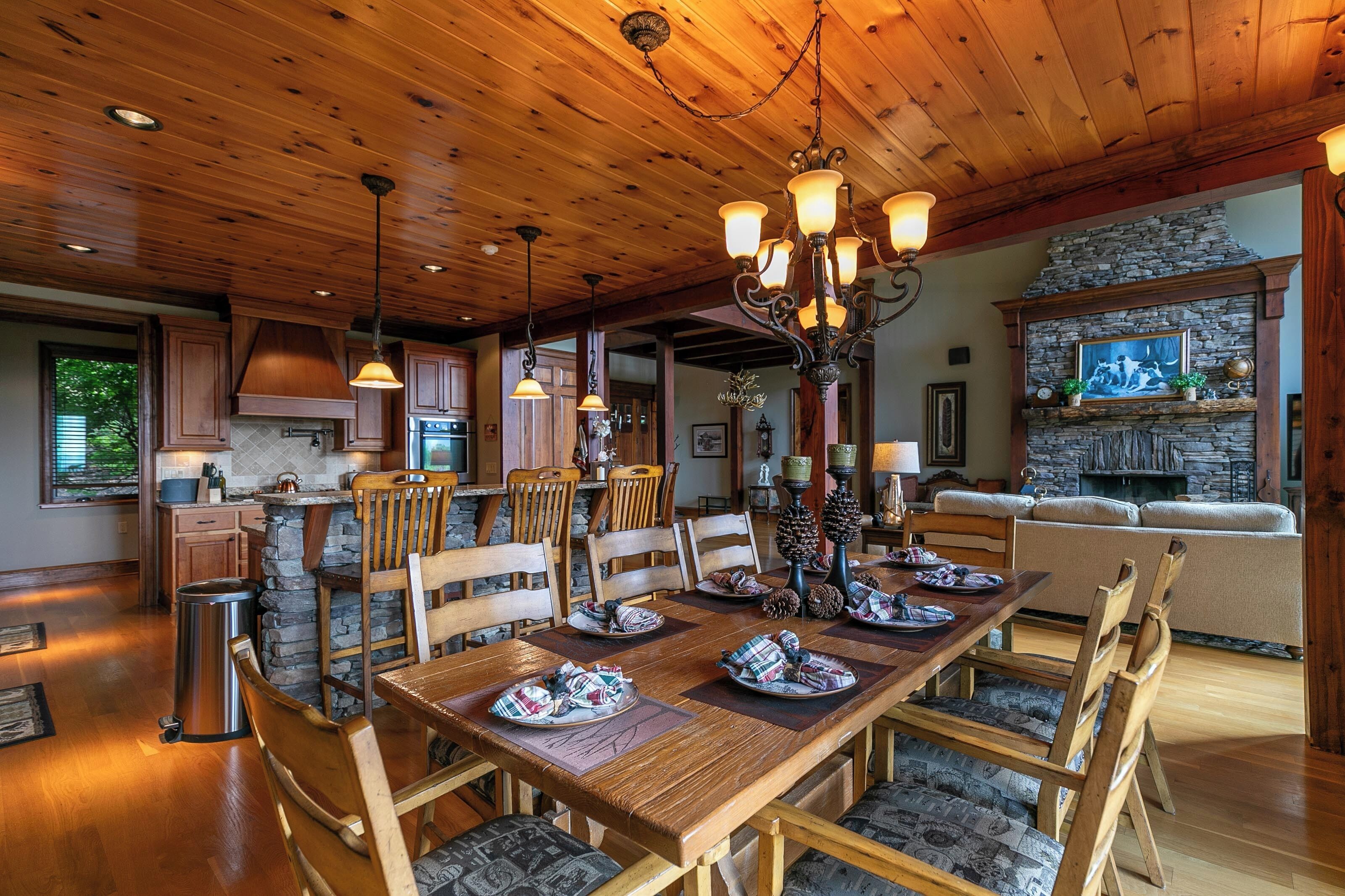 A spacious dining area with a wooden table set for six, surrounded by upholstered chairs. A stone fireplace with a stone mantle and a framed picture above it is visible in the background.