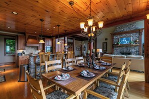 A spacious dining area with a wooden table set for six, surrounded by upholstered chairs. A stone fireplace with a stone mantle and a framed picture above it is visible in the background. - Haven at Headwaters Long-range Mountain Views, Pet Friendly With Game Room (Elk Park)